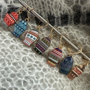 May include: Six colorful mitten-shaped stitch markers with gold rings, hanging on a silver knitting needle. The mittens have different patterns in red, blue, green, and white.