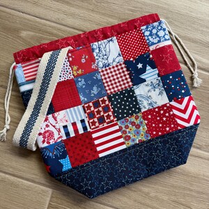 May include: A drawstring bag featuring a patchwork design of red, white, and blue fabric squares with various patterns including stars, stripes, florals, and polka dots. The bag has a dark blue base with stitched stars and a cream-colored strap.