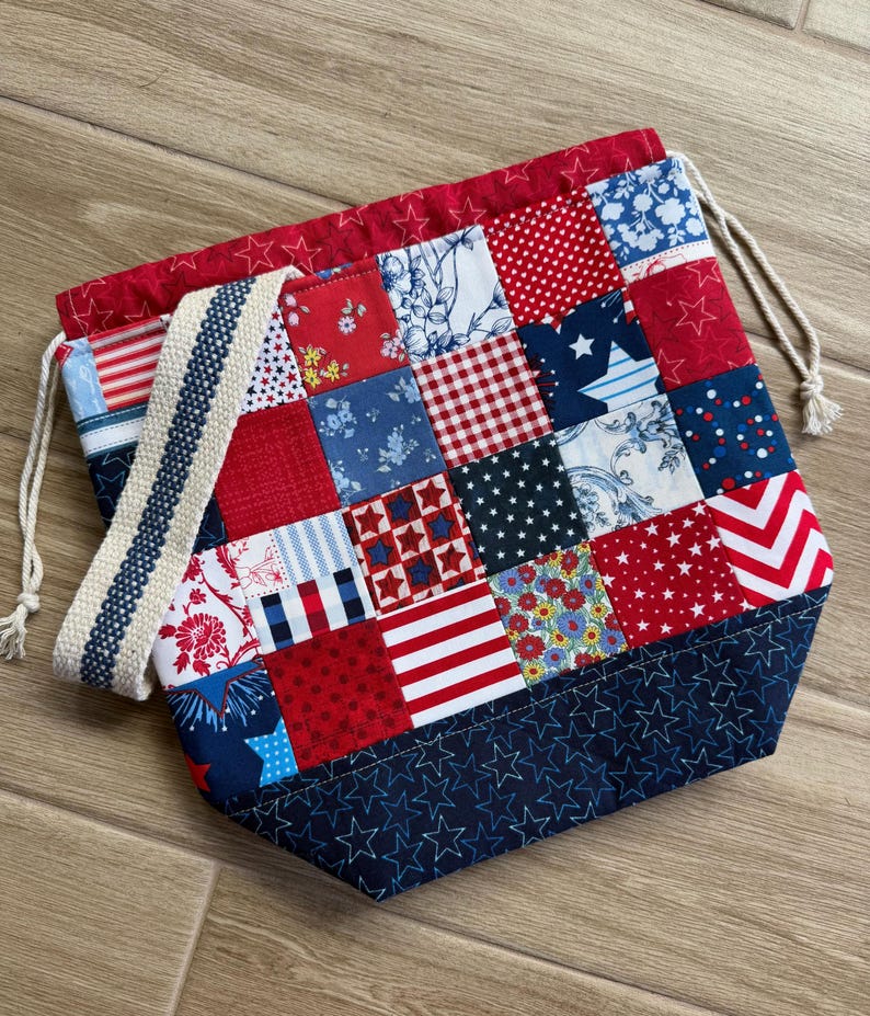 May include: A drawstring bag featuring a patchwork design of red, white, and blue fabric squares with various patterns including stars, stripes, florals, and polka dots. The bag has a dark blue base with stitched stars and a cream-coloured strap.