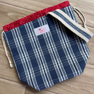 May include: A drawstring bag featuring a navy blue and white plaid pattern on the exterior, a red interior lining with a white star print, and a beige woven strap. A light pink rectangular label with the text Cozy Sky Co is centred.