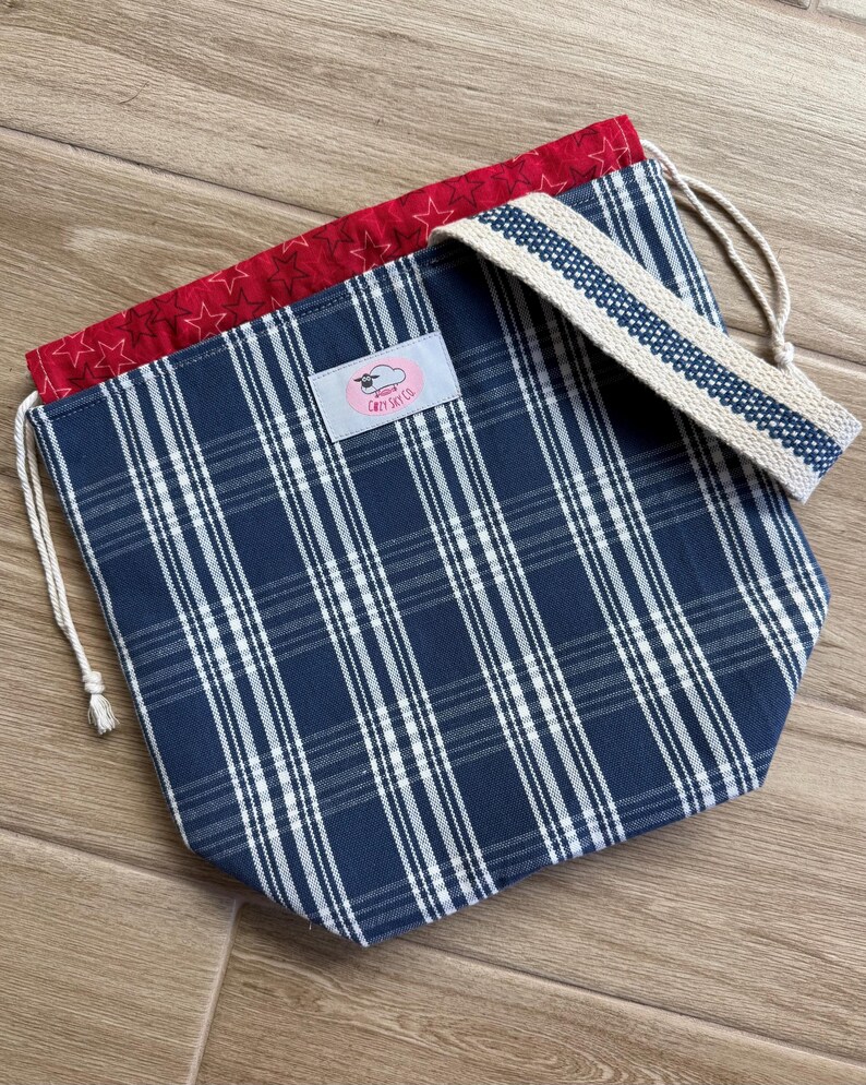May include: A drawstring bag featuring a navy blue and white plaid pattern on the exterior, a red interior lining with a white star print, and a beige woven strap. A light pink rectangular label with the text Cozy Sky Co is centred.