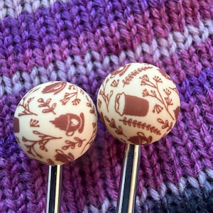 May include: Two ivory-colored knitting needle toppers with brown botanical and coffee-themed designs. The needles are set against a backdrop of purple, lavender, and black knitted fabric, creating a visually appealing contrast.