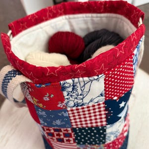 May include: A fabric bucket bag featuring a patchwork exterior with red, white, and blue patterns including stars, gingham, and floral prints. The top edge is trimmed in red fabric with a star pattern, and the bag contains several balls of yarn.