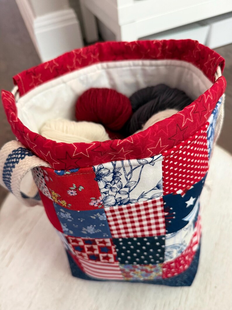 May include: A fabric bucket bag featuring a patchwork exterior with red, white, and blue patterns including stars, gingham, and floral prints. The top edge is trimmed in red fabric with a star pattern, and the bag contains several balls of yarn.