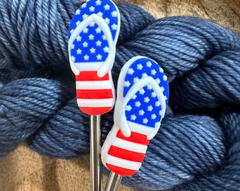 May include: Two silver knitting needle toppers shaped like flip-flops. The flip-flops have a blue star-studded top and red and white striped soles, resembling the American flag. The needles are set against a backdrop of blue yarn.