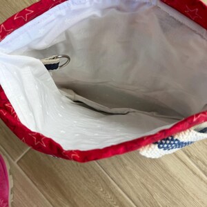 May include: A fabric drawstring bag with a red exterior featuring a white star pattern and a white interior lining. The bag includes a metal D-ring attached to a blue and white woven strap, providing a secure point for closure or attachment.