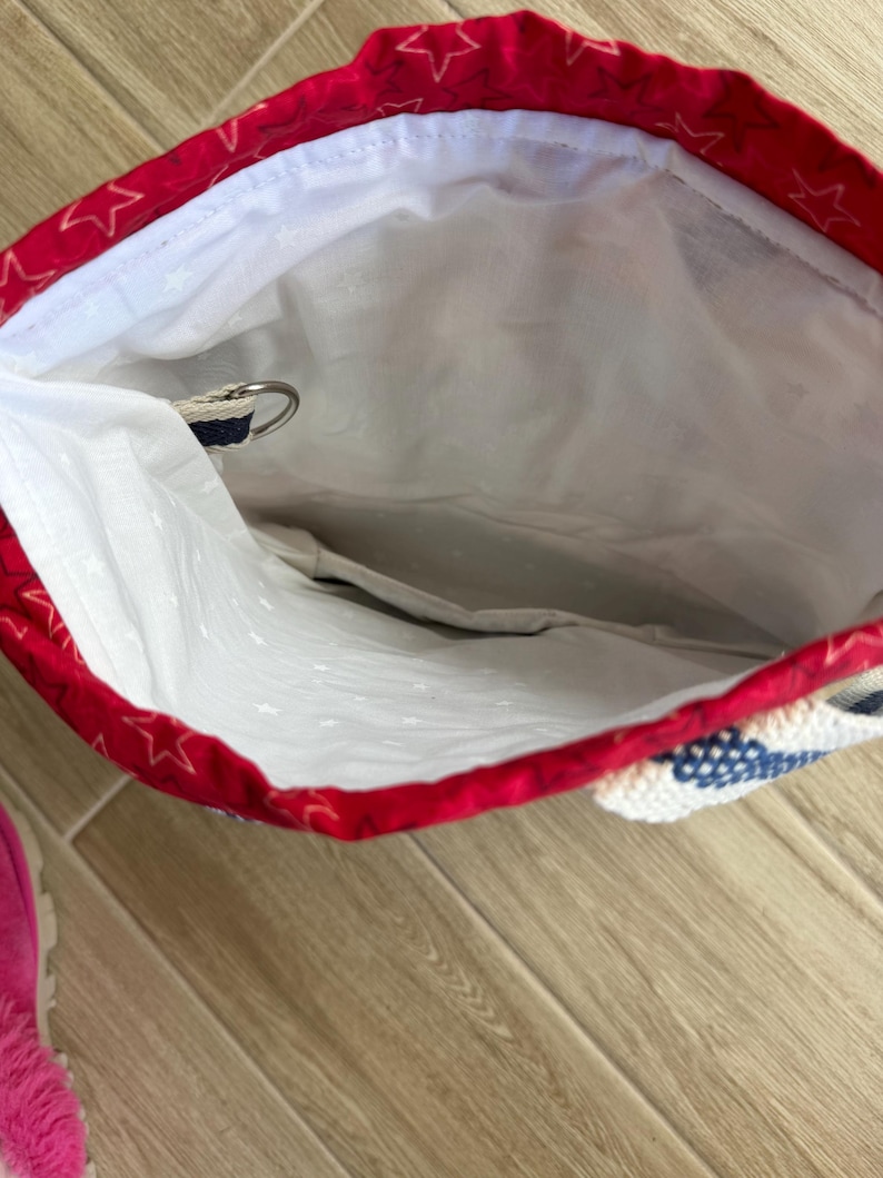 May include: A fabric drawstring bag with a red exterior featuring a white star pattern and a white interior lining. The bag includes a metal D-ring attached to a blue and white woven strap, providing a secure point for closure or attachment.