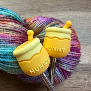 May include: Two silver knitting needles with yellow honey pot shaped ends. The ends have the word "Hunny" written on them. The needles are resting on a ball of colorful yarn.