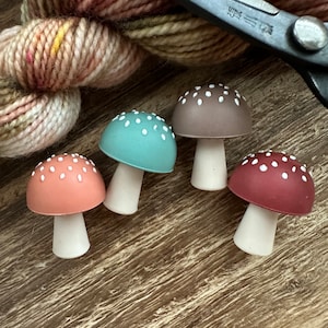 May include: Four miniature mushroom figurines with white polka dots on the caps. The mushrooms are in different colors: teal, brown, coral, and burgundy. The mushrooms are sitting on a wooden surface.