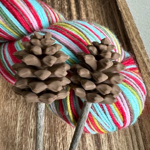 May include: Two brown wooden knitting needles with pine cone-shaped ends. The needles are resting on a ball of colorful yarn with red, pink, blue, green, and yellow stripes.