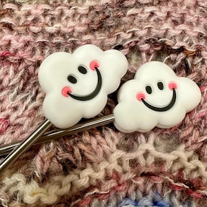 May include: Two silver knitting needles with white cloud-shaped ends. Each cloud has a black smiley face with pink cheeks.