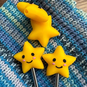 May include: Three yellow star-shaped toppers with smiling faces and silver stems. The toppers are on a blue, gray, and white knitted surface. One star is on its side, and two are upright.