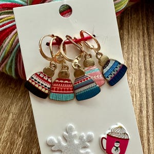 May include: Set of four gold-toned stitch markers with colorful winter hat charms. The charms feature different patterns and colors, including red, blue, and white.