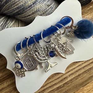 May include: A set of six blue yarn stitch markers with silver charms. The charms include a Star of David, a Hamsa hand, a menorah, an evil eye, and a crescent moon. The stitch markers are displayed on a white card with a blue fuzzy backing.