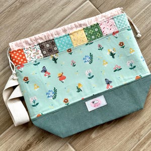 Könnte beinhalten: Ein handgefertigter Beutel mit Kordelzug und einem skurrilen Design. Die Tasche hat einen helltürkisen Stoff, der mit floralen und Insektenillustrationen verziert ist. Der obere Rand hat ein Patchwork-Design mit verschiedenen gemusterten Stoffen. Ein weißer Riemen und Kordelzug vervollständigen die Tasche.