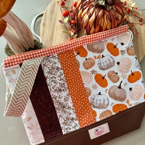 May include: A fall-themed tote bag with a variety of fabrics. The bag features a brown base and a top section with a pumpkin print, houndstooth, floral, and polka dot patterns. The bag is designed with a drawstring closure.