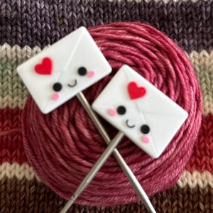 May include: Two knitting needles with white envelope-shaped stitch markers, each featuring a red heart and smiling face, rest on a ball of pink yarn. The background is a striped fabric in shades of pink, brown, and blue.