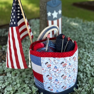 Puede incluir: Un contenedor de almacenamiento de tela rojo, blanco y azul con un tema náutico. El contenedor está lleno de hilo y agujas de tejer. El contenedor está sentado en un lecho de plantas verdes.