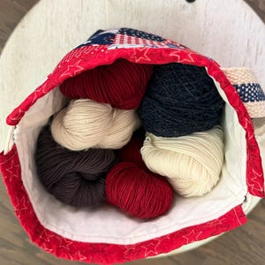 May include: A fabric drawstring bag featuring a red, white, and blue star pattern filled with several skeins of yarn in solid colours including deep red, navy blue, cream, and dark charcoal grey, with a white interior lining and a woven strap.