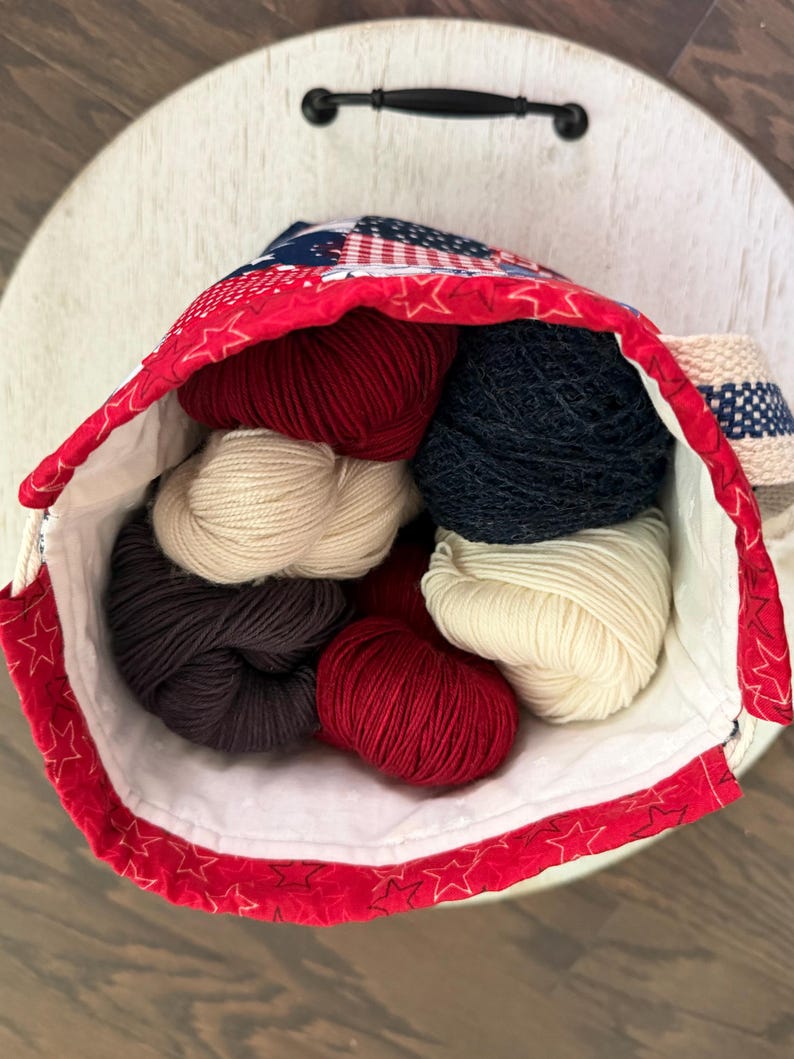 May include: A fabric drawstring bag featuring a red, white, and blue star pattern filled with several skeins of yarn in solid colours including deep red, navy blue, cream, and dark charcoal grey, with a white interior lining and a woven strap.