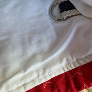 May include: A white fabric lining featuring a subtle pattern of small stars, paired with a red fabric section decorated with dark star outlines. A silver-toned metal D-ring is attached to a navy and white striped fabric strap tucked into the lining.