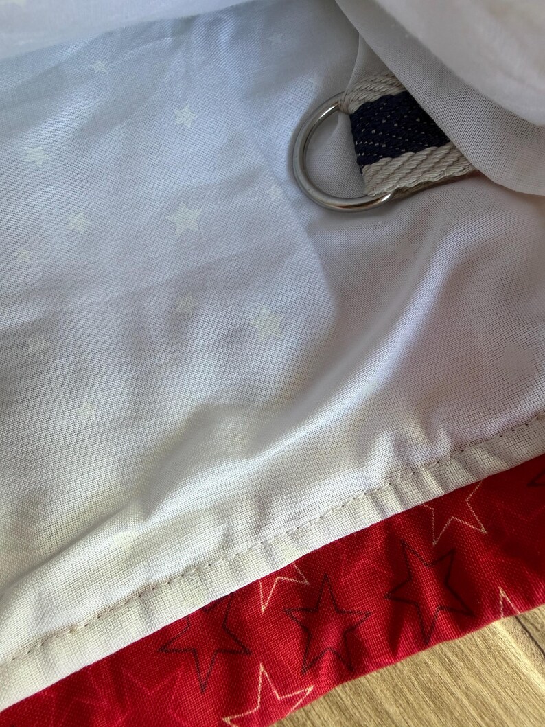 May include: A white fabric lining featuring a subtle pattern of small stars, paired with a red fabric section decorated with dark star outlines. A silver-toned metal D-ring is attached to a navy and white striped fabric strap tucked into the lining.