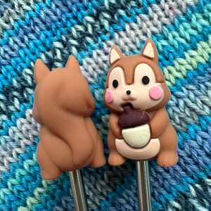 May include: Two knitting needles with brown squirrel-shaped handles. One squirrel is holding a brown acorn. The needles are silver.