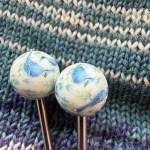 May include: Two knitting needles with round, white tops featuring a blue bird and floral design. The needles are set against a backdrop of knitted fabric in shades of teal, white, and purple.