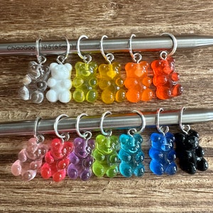 May include: A set of 12 colorful gummy bear stitch markers, each with a silver ring. The markers are arranged in two rows on a wooden surface. The markers are clear, white, yellow, orange, red, pink, purple, green, blue, light blue, dark blue, and black.