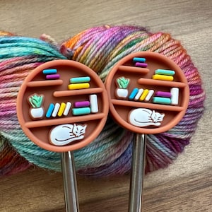 May include: Two orange-brown knitting needle gauges with a bookshelf design. Each gauge features a white sleeping cat, a small plant, and colorful books. The background shows a colorful skein of yarn.