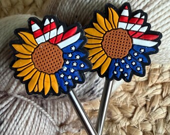 May include: Two metal knitting needles topped with sunflower-shaped charms. The sunflower petals are yellow, while the center is brown. One section of the petals features red and white stripes, and another section is blue with small white stars.