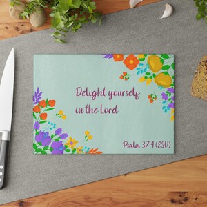 Könnte beinhalten: Ein rechteckiges Glasschneidebrett mit hellblauem Hintergrund, verziert mit bunten Blumenillustrationen in Orange, Gelb und Lila. Der Text lautet "Delight yourself in the Lord" und "Psalm 37:4 (ESV)" in einer dunkelvioletten Schreibschrift.