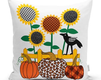May include: A square throw pillow with a white background featuring a graphic illustration of three pumpkins in front of a yellow picket fence. Above the fence are four sunflowers with patterned centers and a black bird with a checkered wing.
