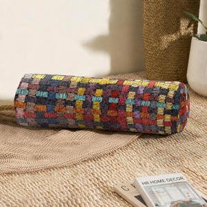 Può includere: Un cuscino cilindrico caratterizzato da un motivo intrecciato con un design a griglia multicolore. Il tessuto presenta una trama ripetitiva di piccoli rettangoli cuciti nelle tonalità del giallo, rosso, blu, arancione e grigio scuro.