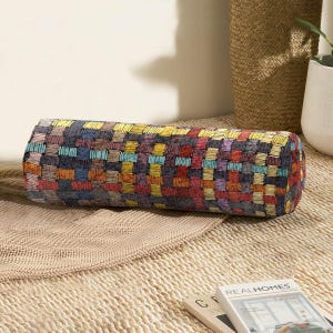 May include: A colourful cylindrical cushion with a geometric pattern of squares in shades of red, yellow, blue, green, and purple. The cushion is made of a soft, textured fabric and is perfect for adding a touch of colour and style to any room.