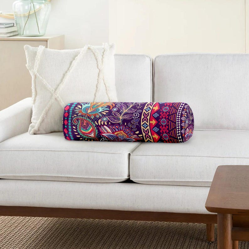 May include: A cylindrical bolster pillow featuring a vibrant purple base with a multi-colored paisley and geometric pattern, positioned alongside a square white throw pillow with a textured, crisscross tufted design on a white sofa.