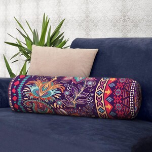 May include: A cylindrical bolster pillow featuring a deep purple base with a vibrant, multi-colored floral and paisley pattern, accented by geometric borders in shades of orange, teal, and pink, resting on a dark blue sofa surface.