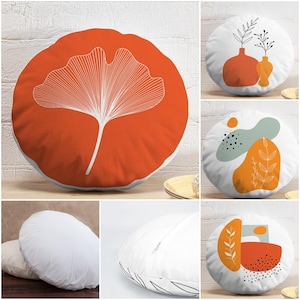 May include: A collection of round decorative cushions. One cushion displays a white ginkgo leaf on an orange backdrop. Other cushions feature abstract designs with orange, teal, and black shapes. The sides of the cushions are white.