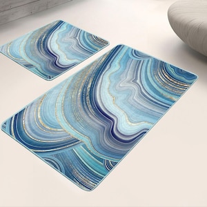 Puede incluir: Dos alfombras decorativas con un diseño de ágata azul y dorado. La alfombra más grande es rectangular y la más pequeña es cuadrada. Las alfombras presentan patrones arremolinados en tonos de azul, verde azulado y dorado, creando un efecto marmolado.