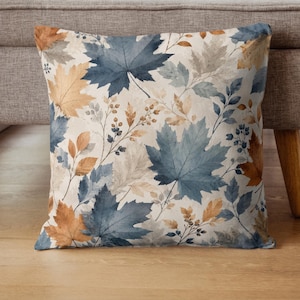 May include: A square throw cushion featuring a repeating pattern of watercolour-style maple leaves and small berries. The design displays a colour palette of muted blue, warm tan, and soft beige tones set against a light cream-coloured background.