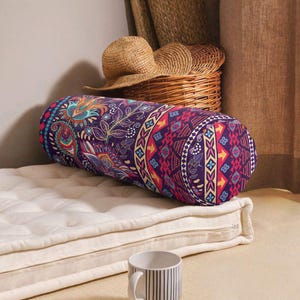 May include: A cylindrical bolster pillow featuring a vibrant purple fabric with a multi-colored paisley and geometric pattern. The pillow is positioned on a white tufted mattress pad, with a straw hat and a woven basket visible in the background.