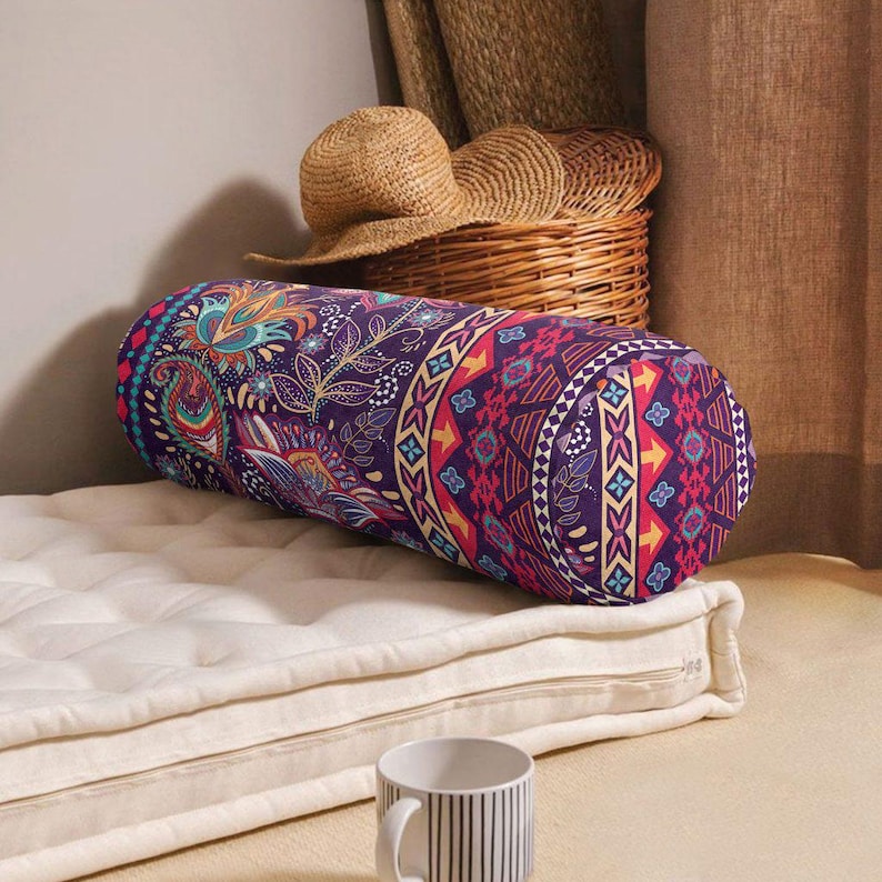 May include: A cylindrical bolster pillow featuring a vibrant purple fabric with a multi-colored paisley and geometric pattern. The pillow is positioned on a white tufted mattress pad, with a straw hat and a woven basket visible in the background.