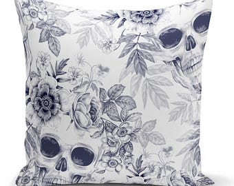 May include: A square throw pillow featuring a white background with a dark blue, detailed botanical illustration of various flowers, leaves, and two human skulls integrated into the floral pattern, creating a high-contrast, intricate design.