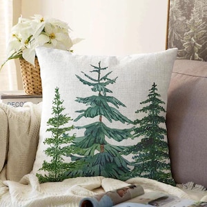 Puede incluir: Un cojín cuadrado con funda de tela blanca que presenta una ilustración impresa de tres árboles de hoja perenne en varios tonos de verde. El árbol central es el más grande, flanqueado por dos árboles más pequeños a cada lado, en estilo acuarela.