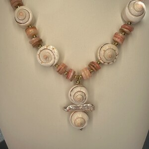 May include: A white shell necklace with pink and gold accents. The necklace features a pendant with three stacked shells and a pearl-like accent.