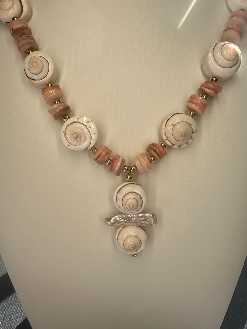 May include: A white shell necklace with pink and gold accents. The necklace features a pendant with three stacked shells and a pearl-like accent.