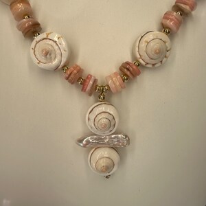 May include: A white seashell necklace with pink and white beads. The necklace has a pendant with three white seashells.