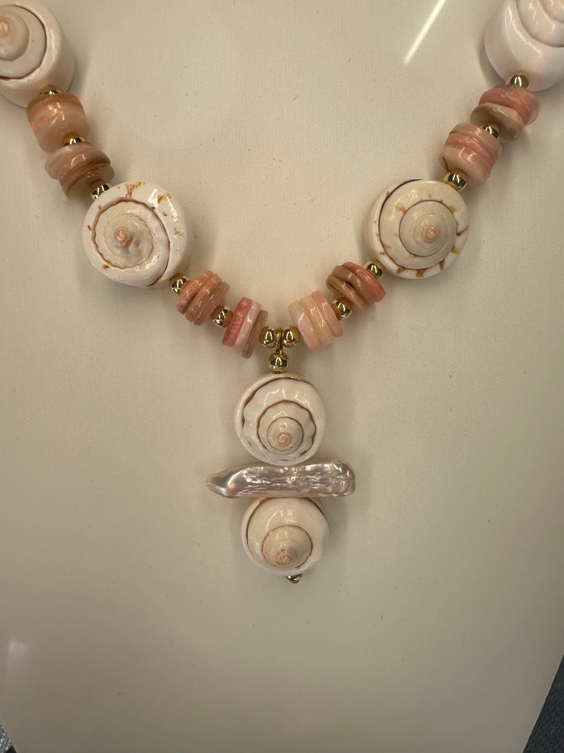 May include: A white seashell necklace with pink and white beads. The necklace has a pendant with three white seashells.