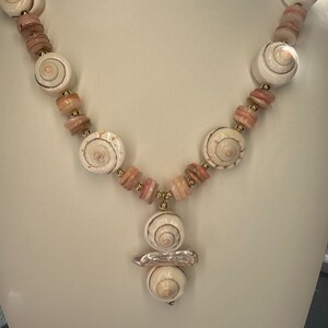 May include: A white and pink shell necklace with a pendant featuring three stacked shells. The necklace is made with small pink and white beads.