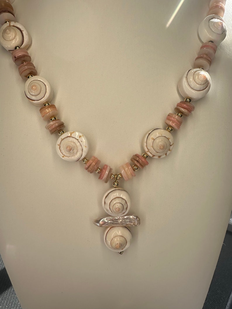 May include: A white and pink shell necklace with a pendant featuring three stacked shells. The necklace is made with small pink and white beads.
